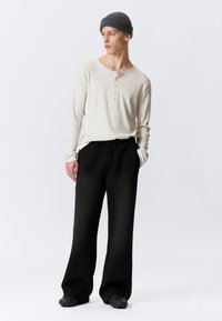 Κρεμ πουκάμισο henley με μακριά μανίκια και κουμπιά, συνοδευόμενο από χαλαρά μαύρα φόρμες και μαύρα παπούτσια με υφή, φορεμένα κάτω από γκρι σκουφί.