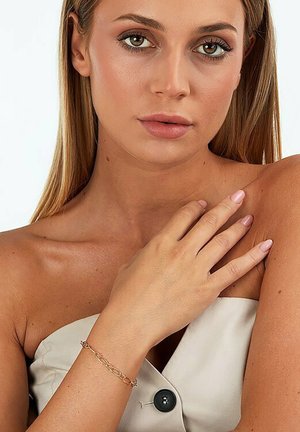 Goldene Kettenarmband mit ovalen Gliedern, das an einem nackten Arm getragen wird, kombiniert mit einem leichten, schulterfreien Oberteil mit einem großen schwarzen Knopfdetail.