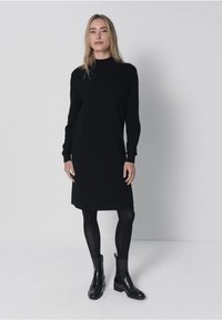 Zwarte jurk met lange mouwen en een hoge halslijn, met een rechte snit en zachte textuur, gecombineerd met zwarte enkellaarzen en panty's.