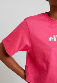 Camiseta corta de algodón rosa con escote redondo y mangas cortas, que presenta texto blanco en la parte delantera. La tela parece suave y ligera.