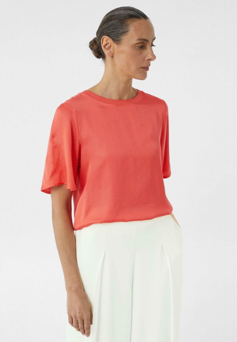 Koraalroze zijden blouse met korte mouwen, ronde halslijn en een ontspannen pasvorm, gecombineerd met witte geplisseerde broeken. Soepele textuur en minimalistisch ontwerp.