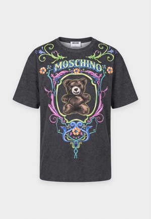 Femme aux cheveux blonds lisses porte un t-shirt Moschino noir avec un motif d'ours en peluche, un pantalon large gris, une ceinture noire et des chaussures noires à bout métallique.