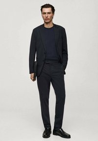 Completo scuro su misura con giacca attillata e pantaloni dritti. Indossato sopra una camicia blu navy, abbinato a scarpe di pelle nere e una pochette.