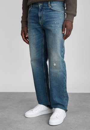 Svetlo modré džínsy s voľným strihom, s vyblednutým umývaním a s odreninami. Skombinované s minimalistickými bielymi teniskami na neutrálnom pozadí.