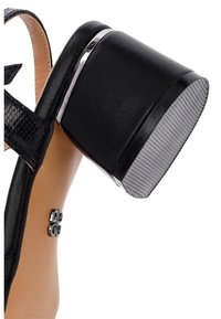 Zwarte sandalen met hakken, gekenmerkt door een chunky hak met een glanzende afwerking, gestructureerde zwarte banden en metallic accenten op de enkelband.