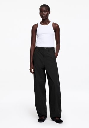 Kobieta stojąca, ubrana w białą bezrękawnikową bluzkę, czarne szerokie spodnie i czarne buty, z jedną ręką w kieszeni na jednolitym białym tle.