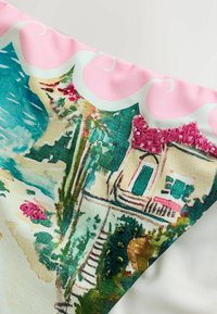 Coin d'écharpe montrant un imprimé coloré de village en bord de mer avec des escaliers, des palmiers et des maisons, accentué d'une broderie perlée rose et verte.