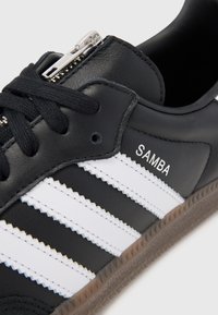 Black leather sneaker with white stripes and "SAMBA" branding. Features a silver zipper and textured brown rubber sole.