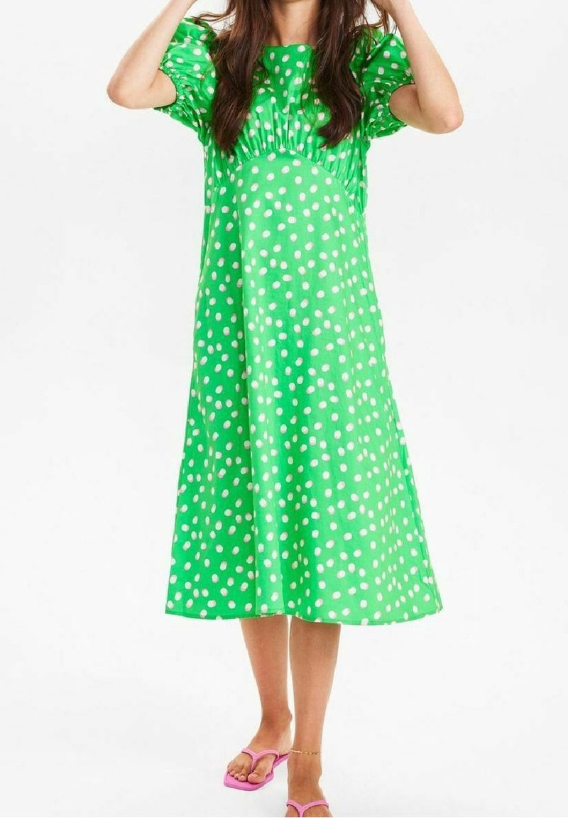 Zielona sukienka w kropki, z krótkimi bufkowymi rękawami, długości do kolan, wykonana z gładkiego materiału. Połączona z różowymi sandałami.