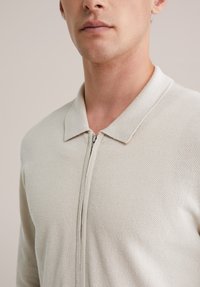 Béžová košeľa na zips s textúrovanou pletenou látkou, s plochým golierom a viditeľným zipsom vpredu, bez ozdôb.