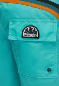 Shorts de bain turquoise avec une poche latérale, comportant un patch logo blanc et marine. Le design inclut des accents orange et noir le long de la taille.