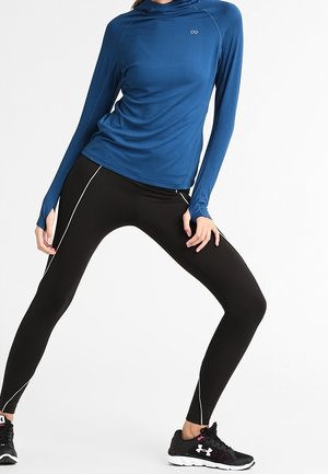 Niebieska sportowa bluzka z długim rękawem o dopasowanym kroju, zestawiona z czarnymi legginsami z białymi paskami po bokach oraz czarnymi butami sportowymi.