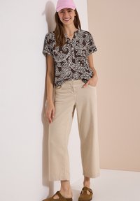 Femme portant une chemise à motif noir et blanc, un pantalon beige à jambes larges, des chaussures marron sans lacets et une casquette de baseball rose, souriante et debout contre un mur.