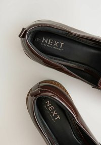 Mocassini in patent marrone con punta arrotondata, soletta nera testurizzata e branding discreto. Presentano una finitura liscia e una suola bassa e resistente.