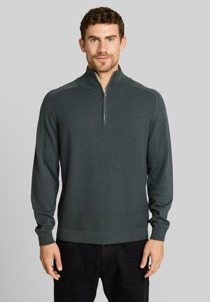 Man met kort krullend haar draagt een donkergrijze trui met rits en zwarte broek, staand tegen een effen lichtgrijze achtergrond.