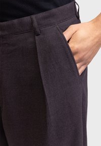 Hand ruht in der vorderen Tasche einer dunkelgrauen Hose aus weichem Stoff, getragen mit schwarzem Oberteil.