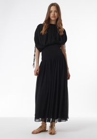 Schwarzes Maxikleid mit einem strukturierten, gerafften Oberteil mit Puffärmeln, einem gerafften Taillenbereich und einem fließenden, durchsichtigen Rock. Goldene Sandalen.