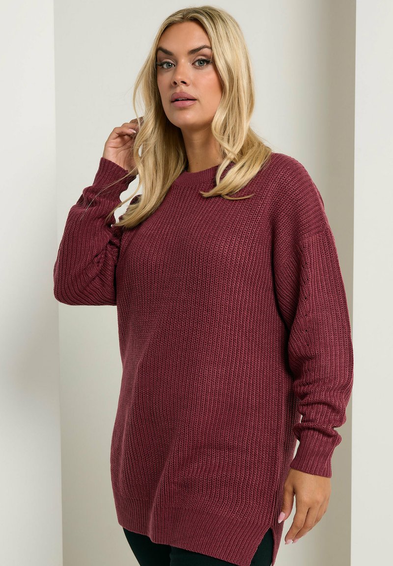 Burgunderfarbener Strickpullover mit lockerer Passform, gerippter Textur, langen Ärmeln, Seitenschlitzen und rundem Halsausschnitt, kombiniert mit schwarzen Leggings.