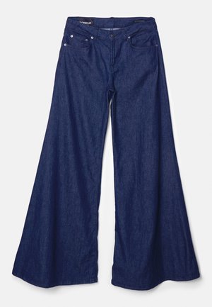 Kvinde iført mørke vidde jeans, sort blazer, hvid top og sorte åbne sko står mod en ensartet grå væg.