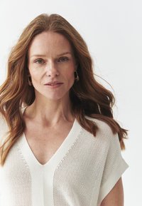Kudottu V-pääntie paita off-white-värissä, jossa on lyhyet hihat ja teksturoitu, avonainen kudontakuvio. Sujuva viimeistely hienovaraisilla yksityiskohdilla.