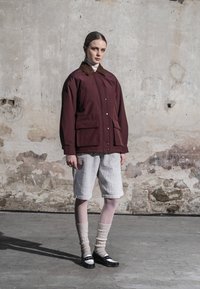 Femme debout contre un mur en béton usé, portant une veste bordeaux, un short blanc côtelé, des chaussettes beiges et des chaussures noires et blanches.