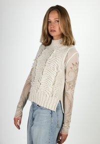 Frau mit schulterlangem Haar trägt einen cremefarbenen, gestrickten ärmellosen Pullover über einer beige-musterten, transparenten Langarmbluse und hellblauen Jeans, steht seitlich.