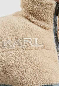 Bež flis jakna s visokim ovratnikom, zatvaranjem na patentni zatvarač i izvezanim logotipom "KARL". Mekana tekstura, ležeran stil i kontrastni plavi detalji.
