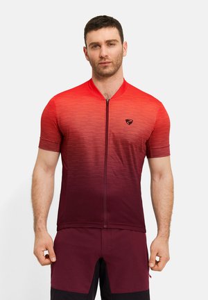 Mann, der ein kurzärmeliges Radtrikot mit einem Farbverlauf von Rot zu Kastanienbraun und eine kastanienbraune Shorts trägt, steht vor einem einfarbigen Hintergrund.