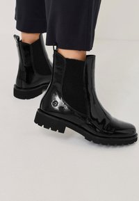 Černé lakované kožené kotníkové boty s elastickými bočními panely, texturovanou černou tkaninou a masivními gumovými podrážkami s vroubkovaným gripem.