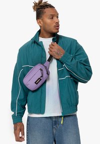 Teal takki tuulenpitävällä, jossa on valkoiset raidat, päällä vaalea T-paita, yhdistettynä purppuraan olkalaukkuun, jossa on vetoketju ja logo-yksityiskohta.