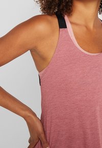 Femme aux cheveux bouclés portant un haut sans manches rose, main posée sur la hanche, montrant le buste contre un fond uni.