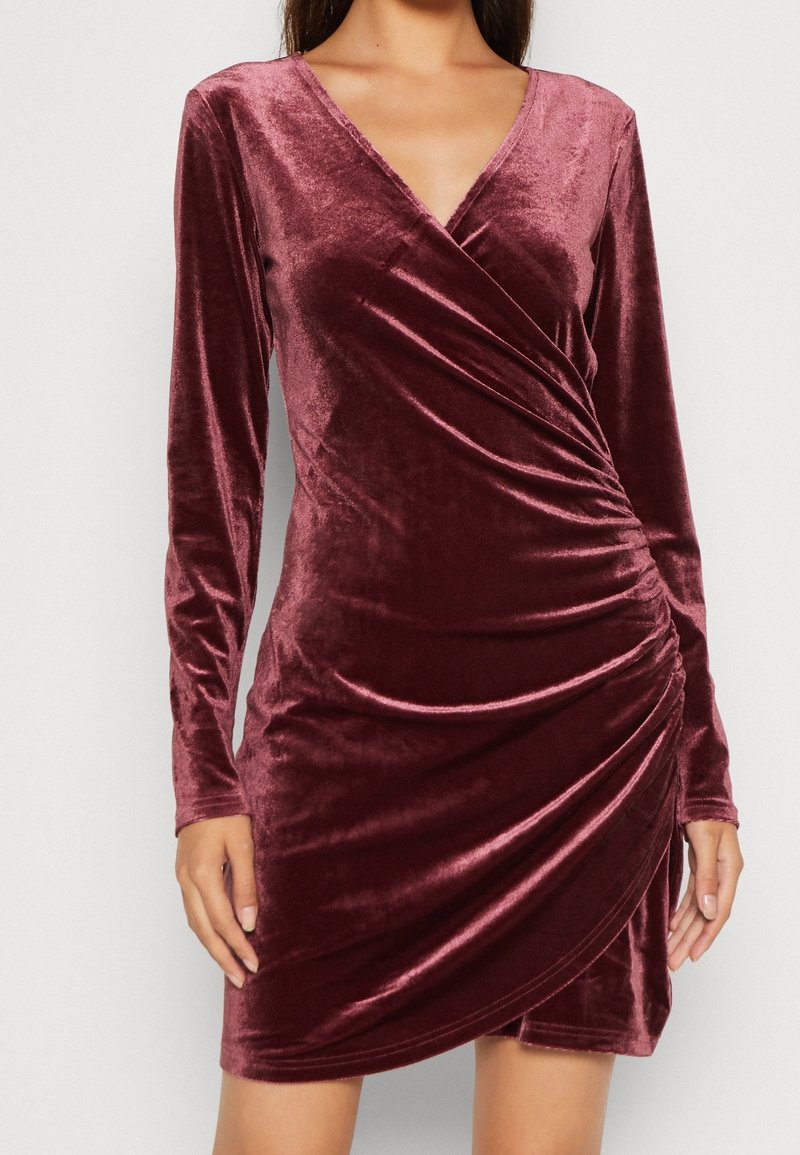 Femme portant une robe portefeuille en velours bordeaux à manches longues, avec détails froncés sur le côté et un col en V.