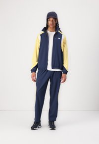 Ensemble de survêtement bleu marine et jaune avec une veste zippée et un pantalon assorti, comprenant des poignets élastiques, un logo sur la poitrine et des panneaux de couleur complémentaire.
