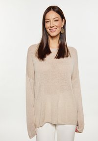 Pulover bej tricotat cu mâneci lungi, gât rotund și croială lejeră. Textura este puțin transparentă, având un aspect moale. Este purtat cu pantaloni albi.