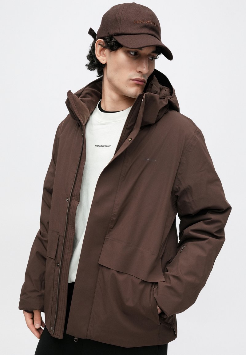 Bruine waterdichte jas met rits- en drukknoopsluitingen, grote voorkantzak en verstelbare capuchon. Gecombineerd met een licht overhemd en bruine pet.