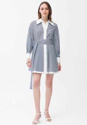 Kobieta ubrana w sukienkę koszulową w pionowe niebiesko-białe paski z szerokim paskiem i białe sandały na wysokim obcasie, stojąca na jednolitym białym tle.