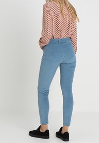 Světle modré úzké sametové kalhoty s zadními kapsami, kombinované s vzorovanou košilí s dlouhým rukávem a černými teniskami.