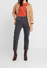 Brązowa kurtka z korduroyu na pomarańczowej koszulce, w połączeniu z czarnymi jeansami o wysokim stanie i brązowymi botkami z wężowej skóry do kostek.