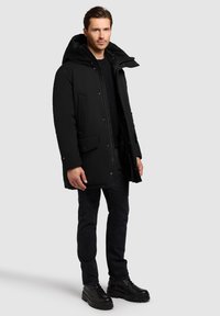 Melna biezināta parka ar kapuci, tai ir divas priekšējās kabatas un sprādzītes. Valkāta kopā ar melnu džemperi, tumšām džinsa biksēm un zābakiem.