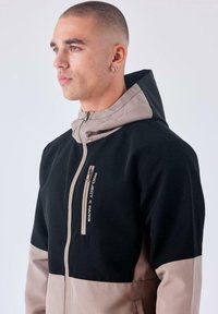 Chaqueta con cremallera negra y beige con capucha, que presenta bloques de color en contraste, una cremallera frontal y detalle del logo "Project X Paris".