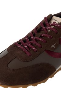 Gros plan sur une basket en daim et cuir marron avec des lacets bordeaux à pois, un logo bordeaux et une semelle en caoutchouc beige.
