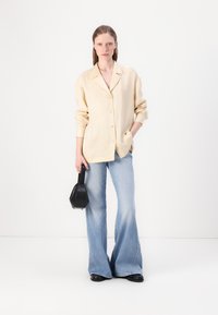 Beige skjorte med knapper og oppbrettede ermer, kombinert med lyseblå flagrende jeans. Holder en liten svart håndveske og står mot en hvit bakgrunn.