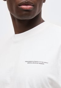 Κοντινό πλάνο ενός ατόμου που φοράει λευκό πουκάμισο με μαύρο κείμενο: «ΑΠΟ ΤΗ FARMÁCIA FRANCO ΣΤΟΝ ΚΟΣΜΟ BENFICA, ΟΔΗΓΗΜΕΝΟ ΑΠΟ ΠΑΘΟΣ.