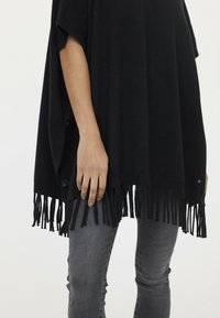 Schwarzer Poncho in lockerer Passform mit fransigem Saum und seitlichen Knopfdetails. Über grauen Jeans getragen. Weicher Stoff.