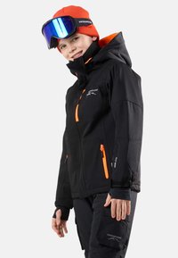 Svart skidjacka med orange detaljer, utrustad med huva, dragkedjefickor och justerbara ärmar. Bärs med en orange mössa och skidglasögon.