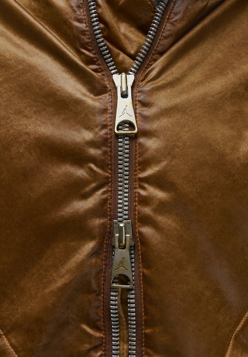 Braune Lederjacke mit glänzender Oberfläche, ausgestattet mit zwei silberfarbenen Reißverschlüssen und einem auffälligen Logo-Akzent in der Mitte.