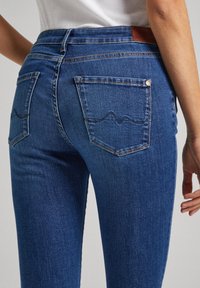 Jeans skinny de color azul oscuro con una textura de mezclilla suave, que cuentan con dos bolsillos traseros con un patrón de costura en zigzag y una etiqueta de cuero marrón en la cintura.