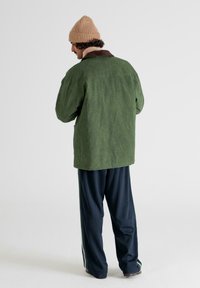 Grøn, struktureret jakke med en brun krave, parret med mørke bukser med side-striber og brune sko. Modellen bærer en beige strikket hue.