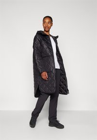 Black quilted long puffer coat with a hood, featuring diagonal stitching, side pockets, worn over a white shirt and black pants.