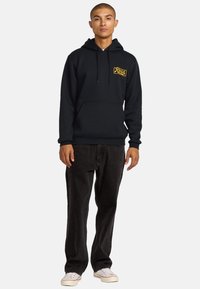 Black hoodie with a front pocket, features a yellow logo on the left chest. Paired with dark denim jeans and white sneakers.
